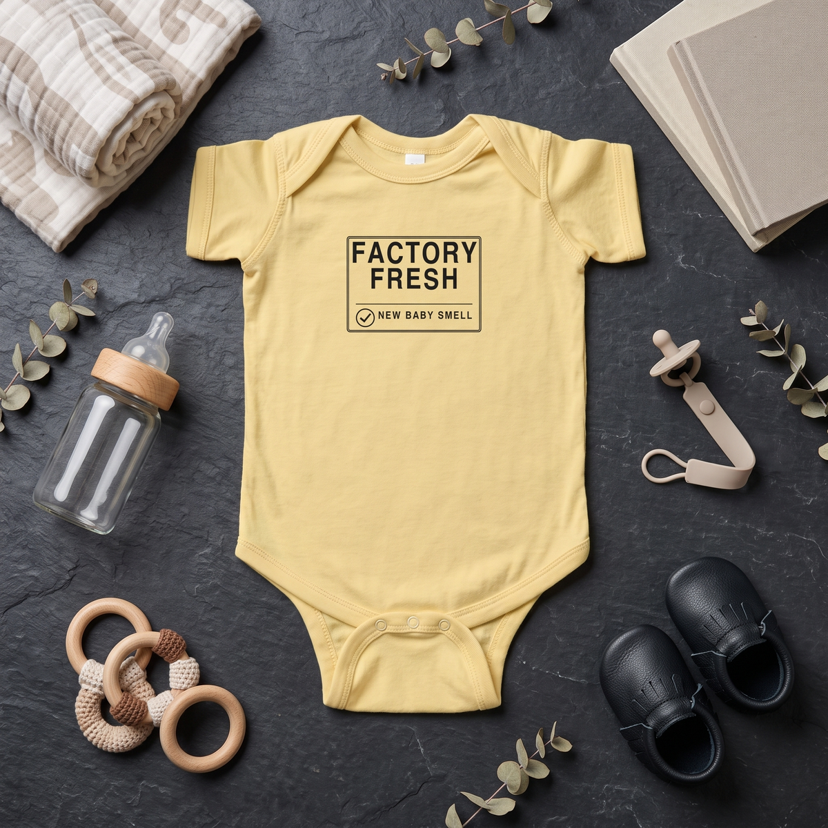 A pale yellow baby bodysuit laid flat on a dark textured surface, surrounded by baby-themed props including a bottle, pacifier, shoes, rattle, blanket, and books. The bodysuit has a black rectangular chest print reading “FACTORY FRESH” and “NEW BABY SMELL.