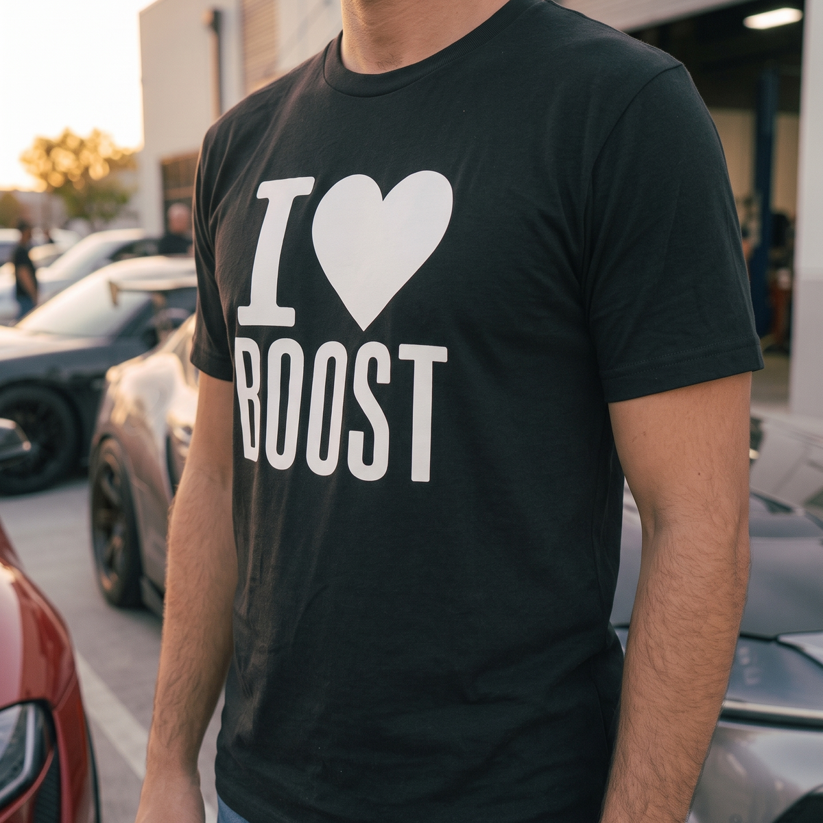 A man wearing a black crewneck t-shirt with a bold white graphic that reads 'I [heart] BOOST'. The photo is taken outdoors in a parking lot with cars and a garage in the background.