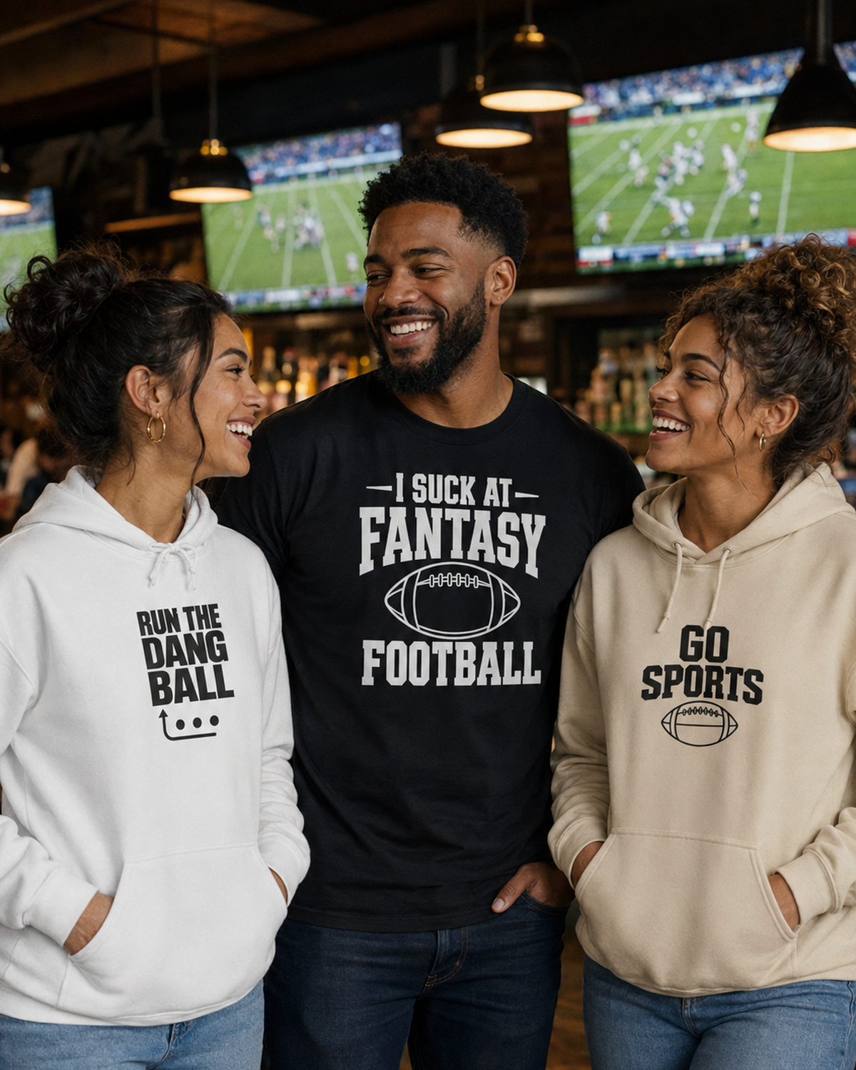 Three friends in a bar or sports bar setting, wearing humorous graphic apparel. The man in the center wears a black t-shirt that reads 'I SUCK AT FANTASY FOOTBALL' with a football graphic.
